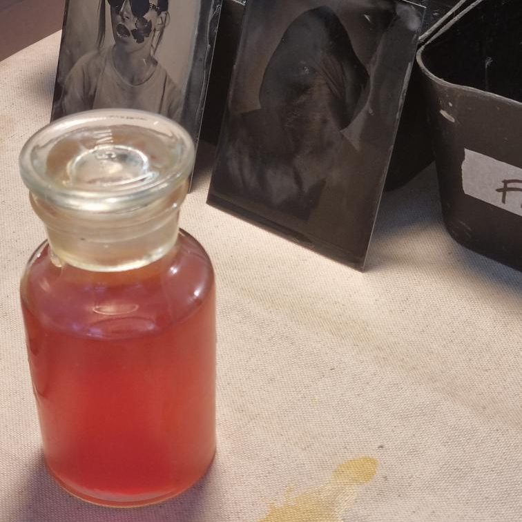 Photo d'une flasque de solution de collodion salé rouge posée devant deux plaque de portraits en train de sécher