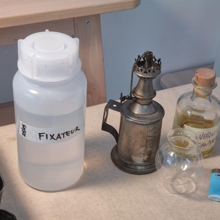Photo d'une lampe à alcool à coté d'une bouteille, contenant un liquide transparent, avec une étiquette marquée "fixateur"